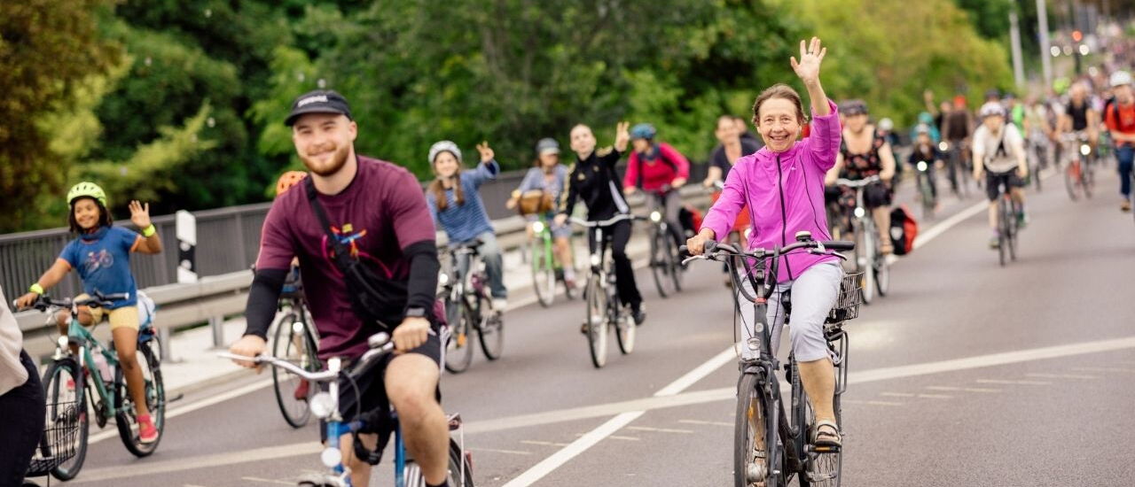 Gruppe von Radfahrern auf einer Straße, vorne eine Frau in pinker Jacke winkt und ein Mann mit schwarzem Pullover und Mütze fährt neben ihr
