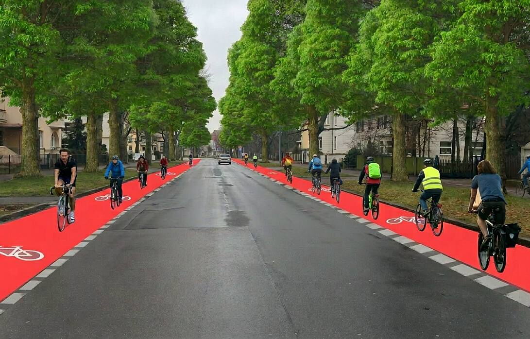 Eine breite, asphaltierte Straße wird von hohen, belaubten Bäumen gesäumt. Auf beiden Seiten der Fahrbahn verlaufen auffällig rote Radwege mit weißen Fahrradsymbolen.