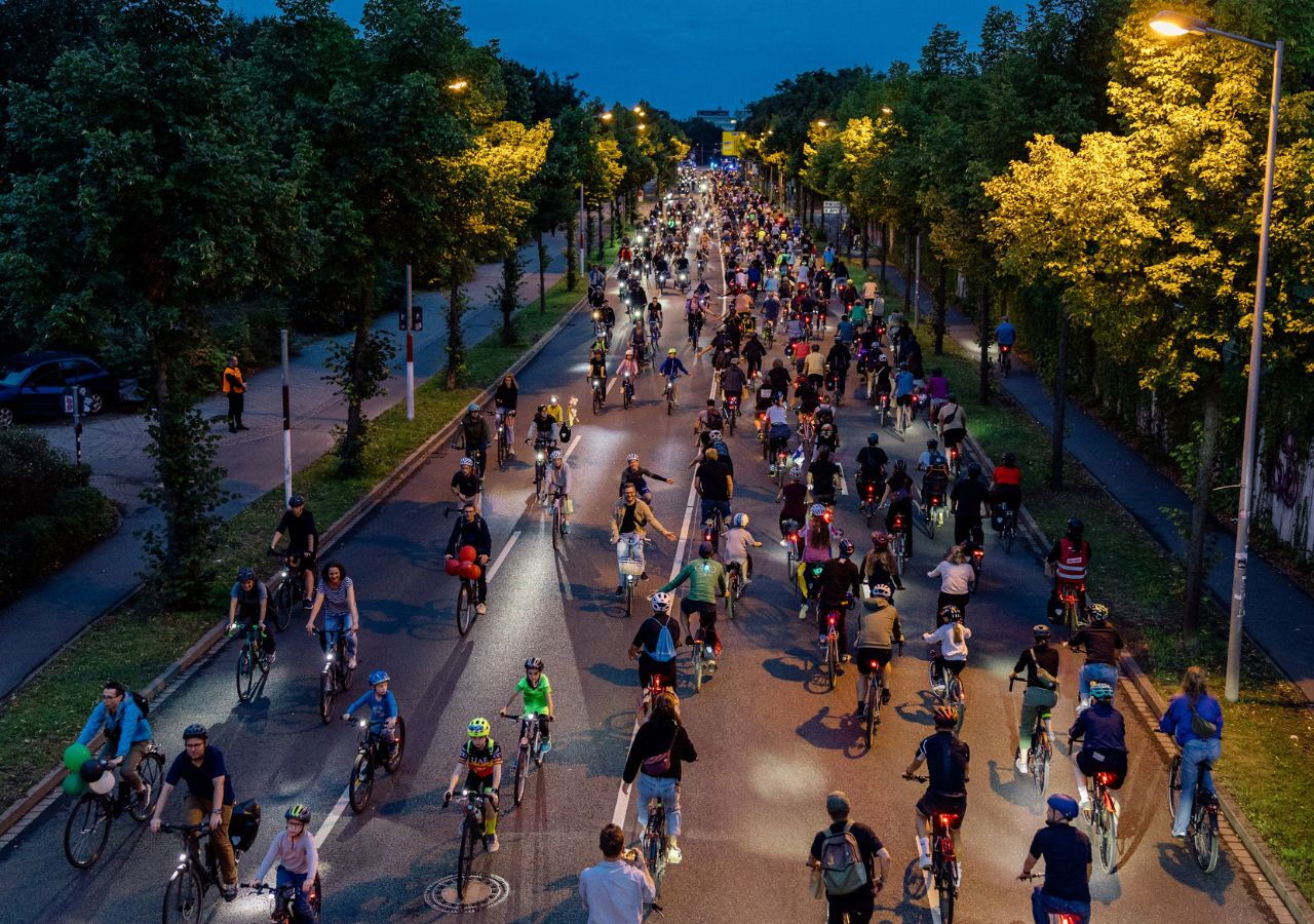 Große Gruppe von Radfahrern auf einer breiten Straße bei Dämmerung umgeben von Bäumen und Straßenlaternen