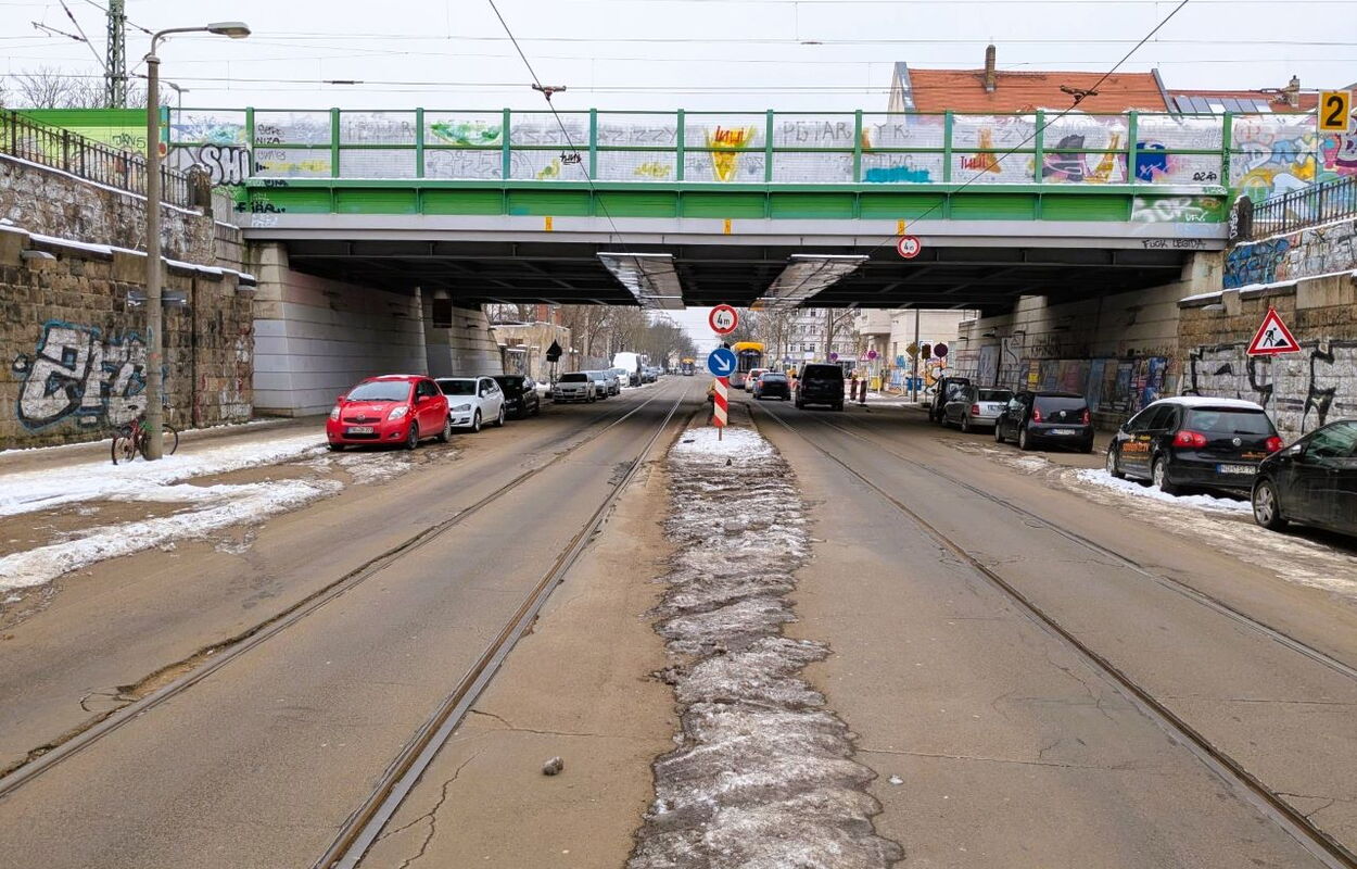 Straße mit Straßenbahnschienen unter einer Brücke mit Graffiti an den Brückenwänden und mehreren parkenden Autos auf beiden Seiten