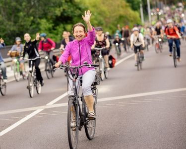 Frau in pinker Jacke winkt lächelnd auf Fahrrad in einer Gruppe von Radfahrern auf Straße