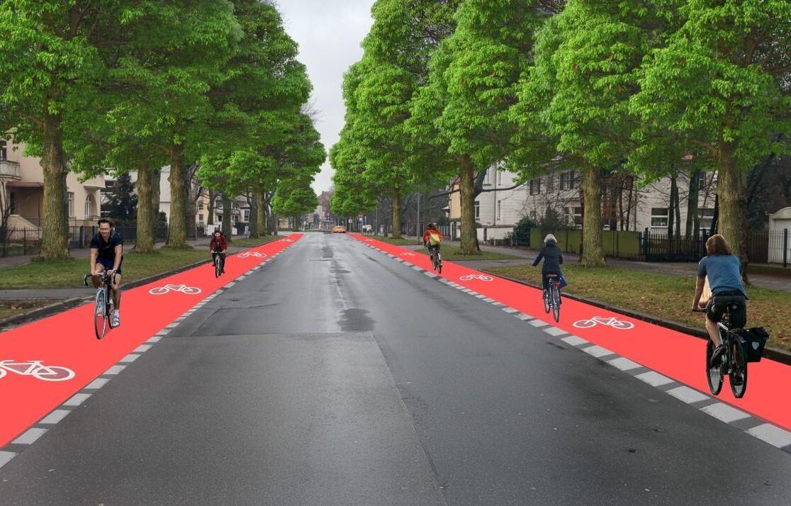 Eine breite, asphaltierte Straße wird von hohen, belaubten Bäumen gesäumt. Auf beiden Seiten der Fahrbahn verlaufen auffällig rote Radwege mit weißen Fahrradsymbolen.
