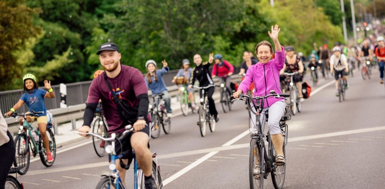 Gruppe von Radfahrern auf einer Straße, vorne eine Frau in pinker Jacke winkt und ein Mann mit schwarzem Pullover und Mütze fährt neben ihr