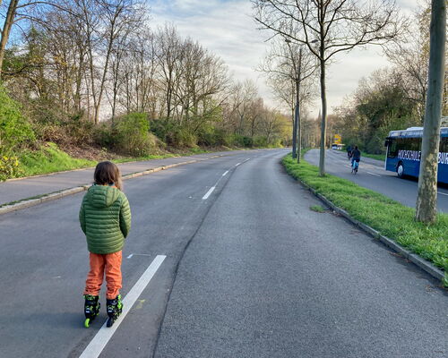Kind mit grüner Jacke und oranger Hose fährt Inliner auf einer leeren Straße mit Bäumen und einem Bus im Hintergrund