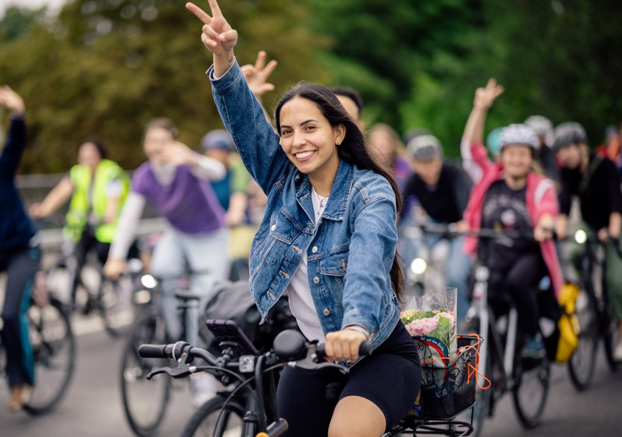Junge Frau auf Fahrrad zeigt lachend Victory-Zeichen, im Hintergrund weitere Radfahrer in lockerer Gruppe auf Straße