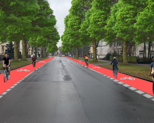 Eine breite, asphaltierte Straße wird von hohen, belaubten Bäumen gesäumt. Auf beiden Seiten der Fahrbahn verlaufen auffällig rote Radwege mit weißen Fahrradsymbolen.