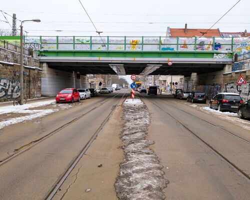 Straße mit Straßenbahnschienen unter einer Brücke mit Graffiti an den Brückenwänden und mehreren parkenden Autos auf beiden Seiten
