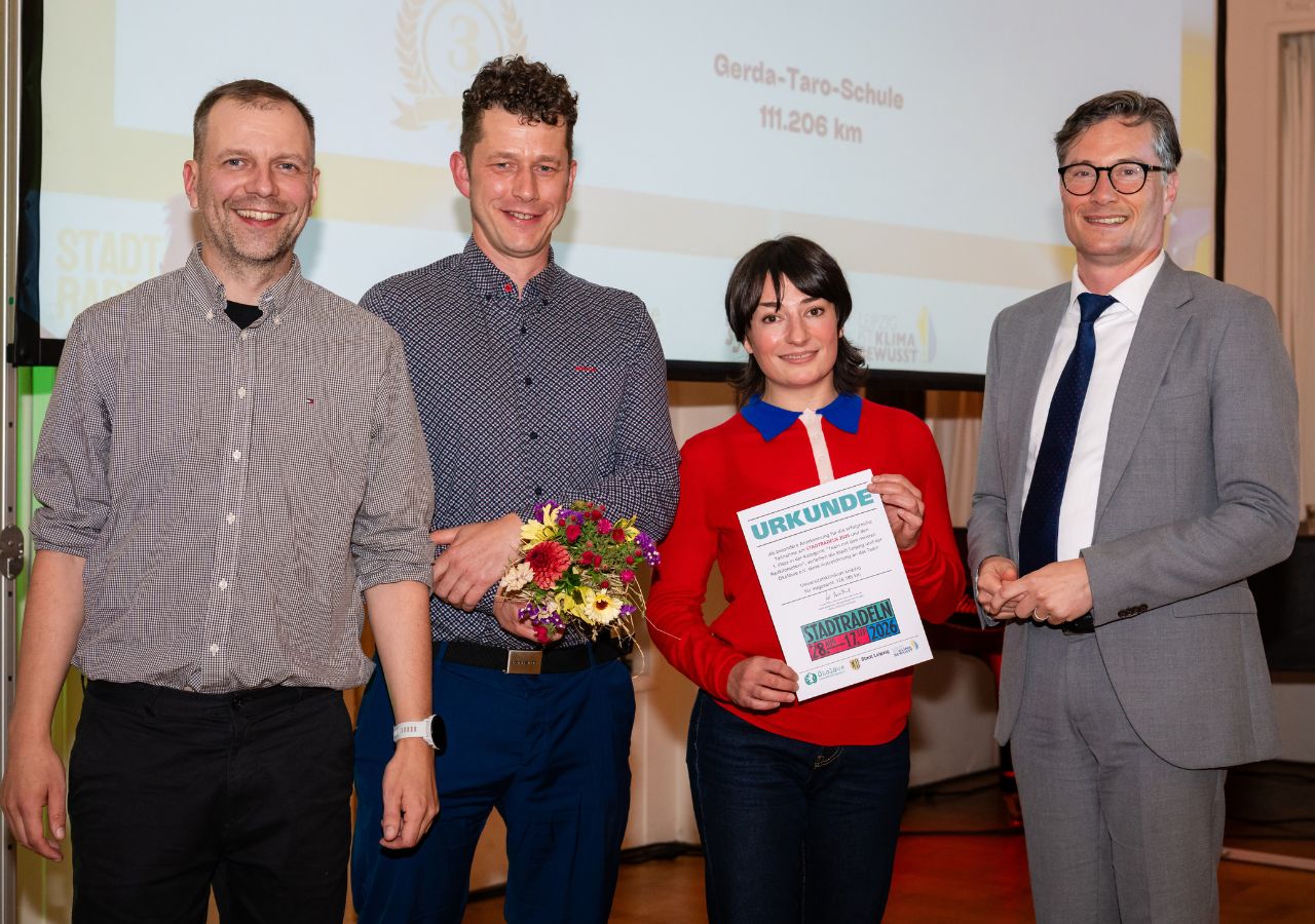 Vier Personen stehen nebeneinander, eine Frau hält eine Urkunde, ein Mann hält einen Blumenstrauß, alle lächeln, im Hintergrund eine Projektionswand