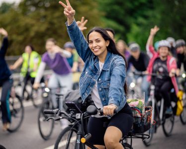 Junge Frau auf Fahrrad zeigt lachend Victory-Zeichen, im Hintergrund weitere Radfahrer in lockerer Gruppe auf Straße