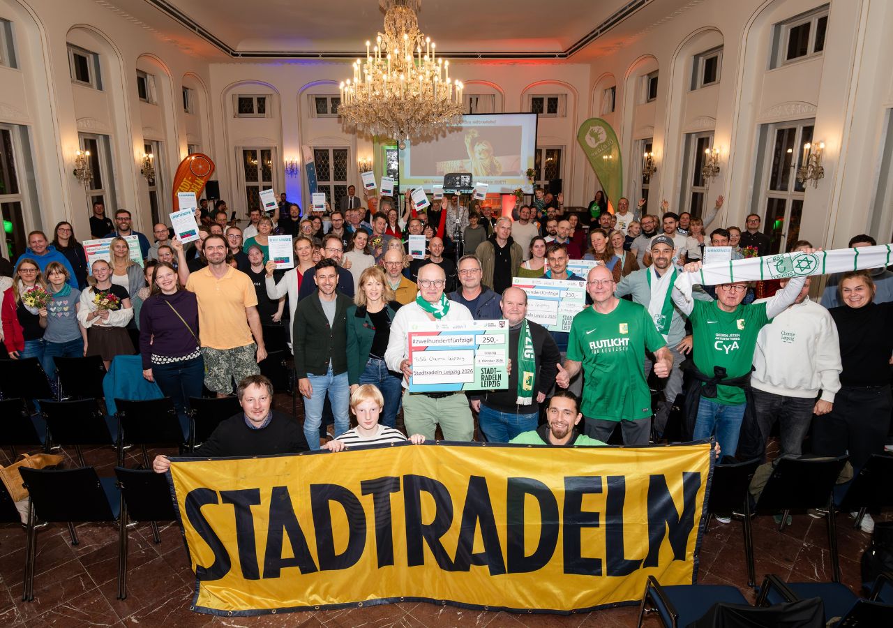 Große Gruppe von Menschen in einem Saal, einige halten Urkunden und einen großen gelben Banner mit der Aufschrift 'STADTRADELN'