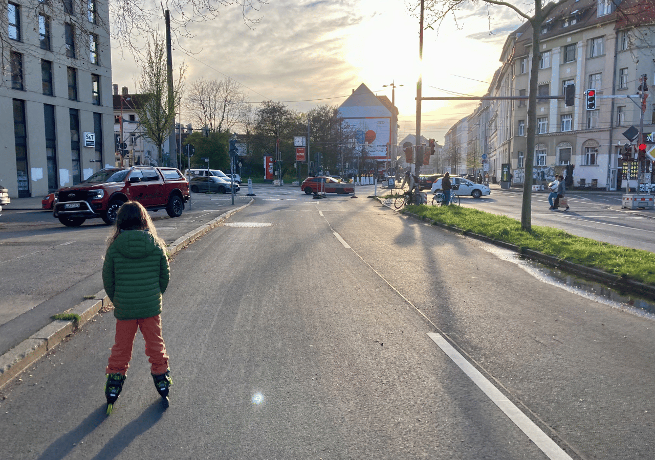 Kind mit grüner Jacke und oranger Hose auf Inlinern steht auf einer Straße vor einer Kreuzung in einer Stadt bei Sonnenuntergang