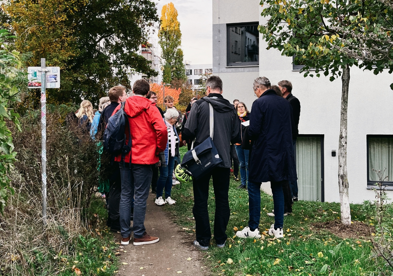 Gruppe von Menschen steht auf einem schmalen Weg neben einem weißen Gebäude, umgeben von Bäumen mit herbstlichen Blättern.