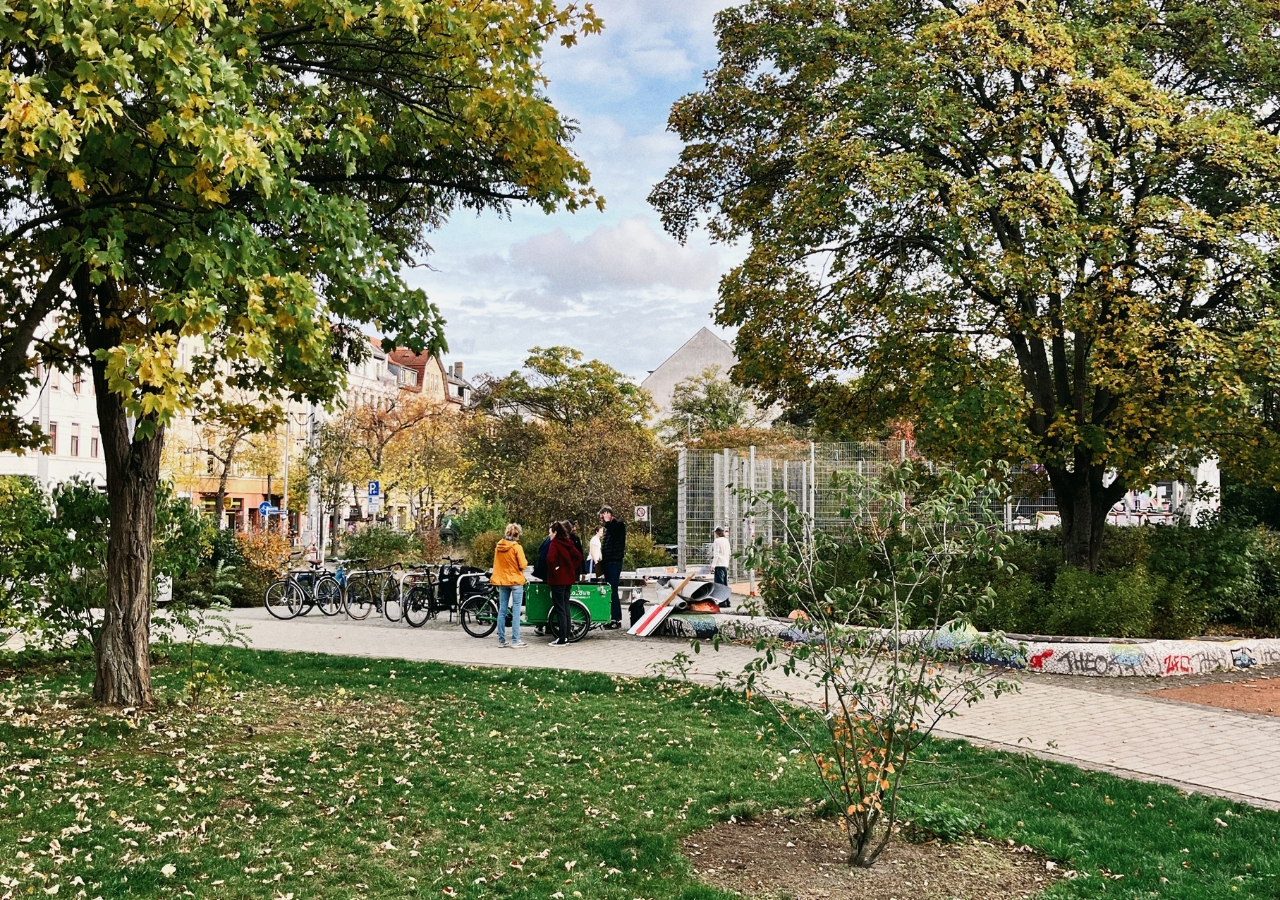 Park mit Bäumen und Rasen, drei Personen stehen an einem Fahrradständer, im Hintergrund Wohnhäuser und blauer Himmel mit Wolken.