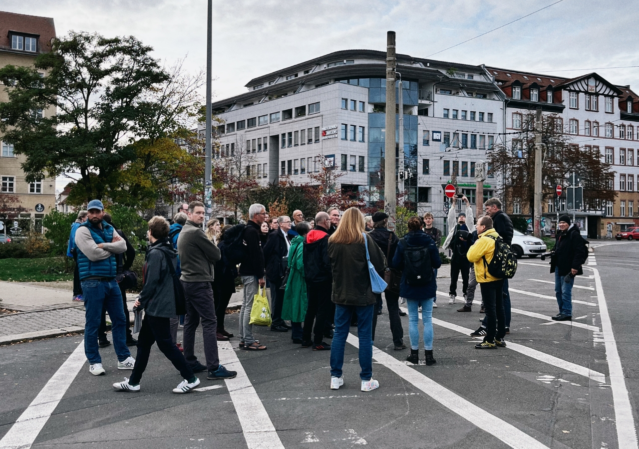 Gruppe von Menschen steht auf einer Kreuzung in einer Stadt mit mehrstöckigen Gebäuden im Hintergrund.