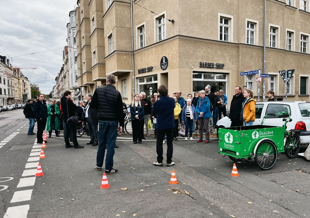 Menschen stehen auf einer Straßenecke vor einem Gebäude mit Schildern 'BARBER SHOP' und 'FRISEUR'. Ein grünes Lastenfahrrad steht am Straßenrand. Der Gehweg ist mit roten Hütchen zu einer Gehwegnase vergrößert