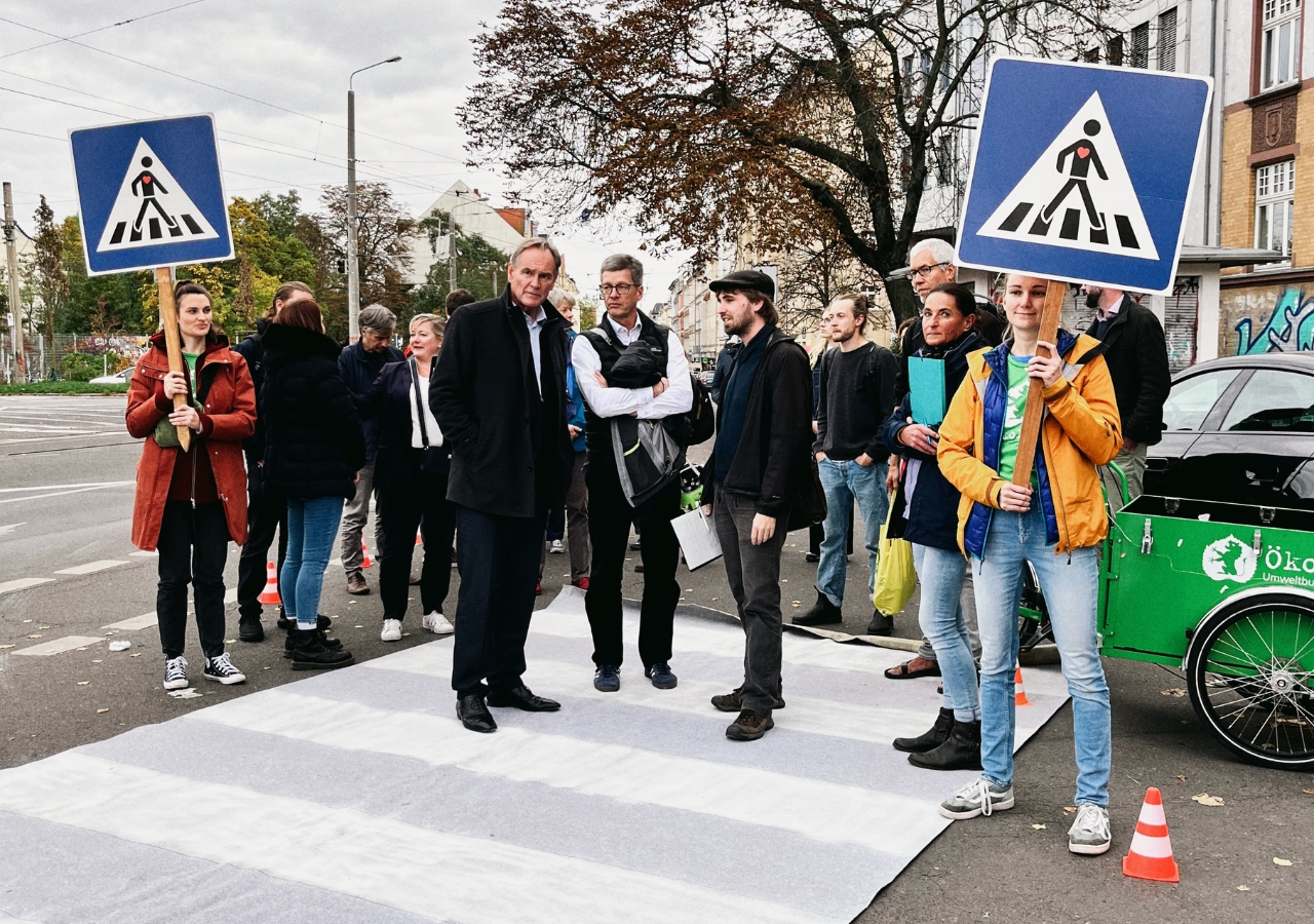 Menschen stehen auf einem ausgerollten Zebrastreifen-Teppich, zwei halten Verkehrsschilder mit Fußgänger-Symbolen, im Hintergrund Bäume und Gebäude.