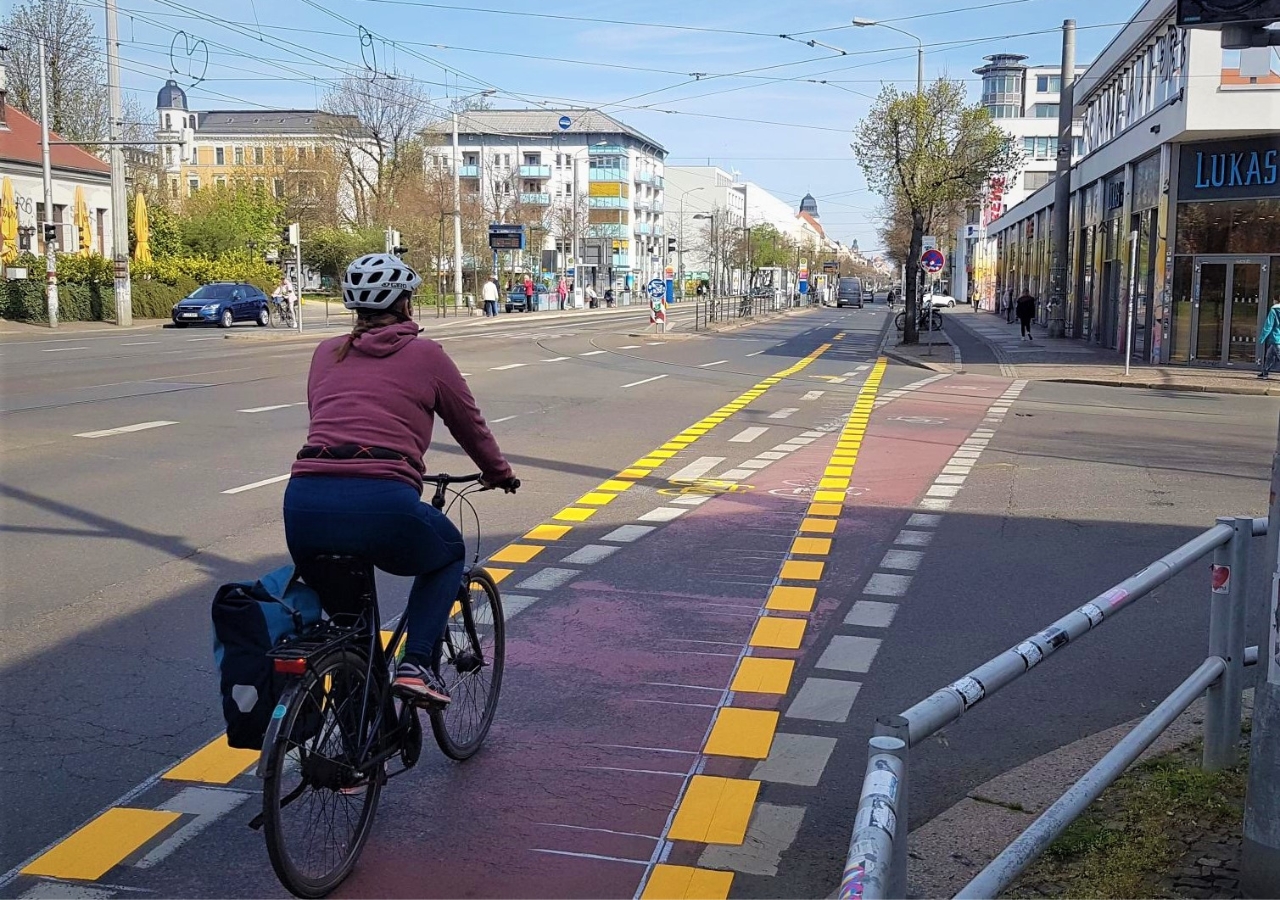 Fahrradfahrerin mit Helm auf rotem Radweg mit gelben Markierungen an einer Straße mit Gebäuden und Bäumen im Hintergrund.