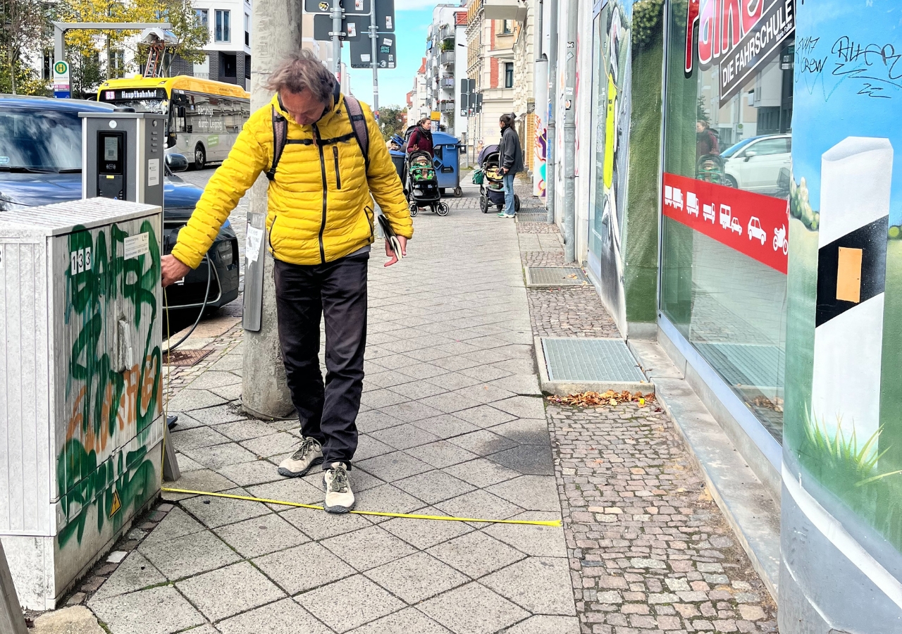 Person mit gelber Jacke und Rucksack vermisst mit einem Maßband die Breite des Gehwegs zwischen Hauswand, abgesenkter Bordsteinkante. und Ladesäule.