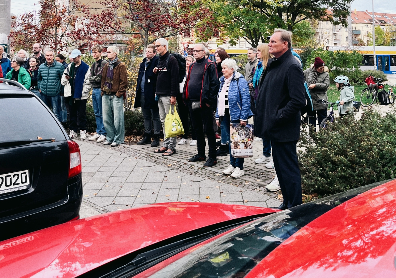 Menschen stehen auf einem Gehweg neben einer Straße mit parkenden Autos, im Hintergrund Bäume und ein Fahrrad.