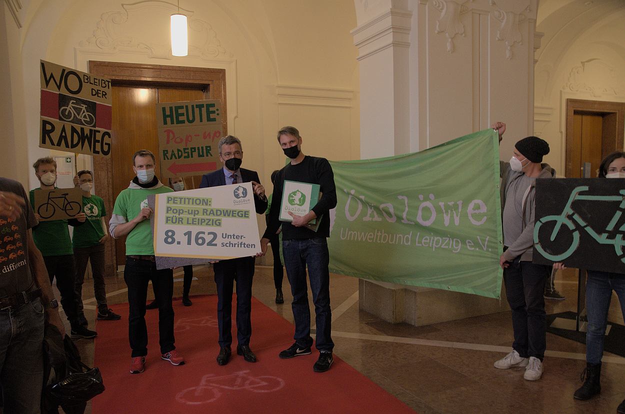 Petitionsübergabe im Neuen Rathaus an Bürgermeister Thomas Dienberg. Schild mit der Aufschrift 'PETITION: POP-UP RADWEGE FÜR LEIPZIG 8.162 Unterschriften', eine weitere Person hält ein grünes Banner mit dem Schriftzug "Ökolöwe"