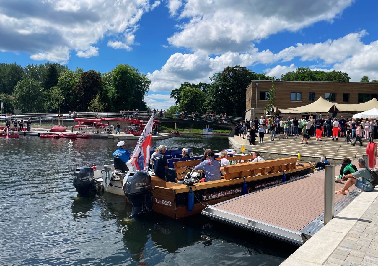 Mehrere Personen sitzen auf einem Holzboot im Stadthafen, im Hintergrund weitere Boote und Bäume