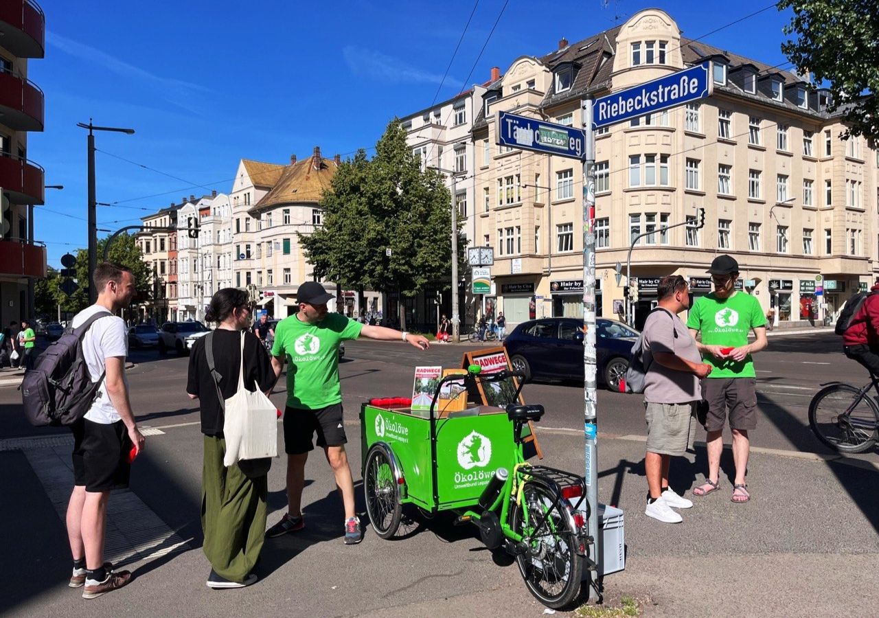 Zwei Männer in grünen T-Shirts mit Logo stehen an einer Kreuzung neben einem grünen Lastenfahrrad mit Aufschrift "ÖkoLöwe" unter Straßenschildern "Riebeckstraße" und "Täubchenweg"