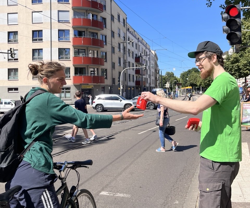 Zwei Personen auf einer Straße, eine reicht der anderen einen Verbandskasten, im Hintergrund Wohngebäude und ein Fahrrad