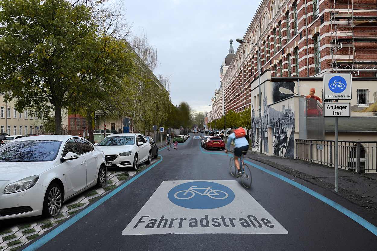 Straße mit blau markiertem Fahrradweg und weißer Aufschrift 'Fahrradstraße', Radfahrer und parkende Autos an der Seite, großes Gebäude